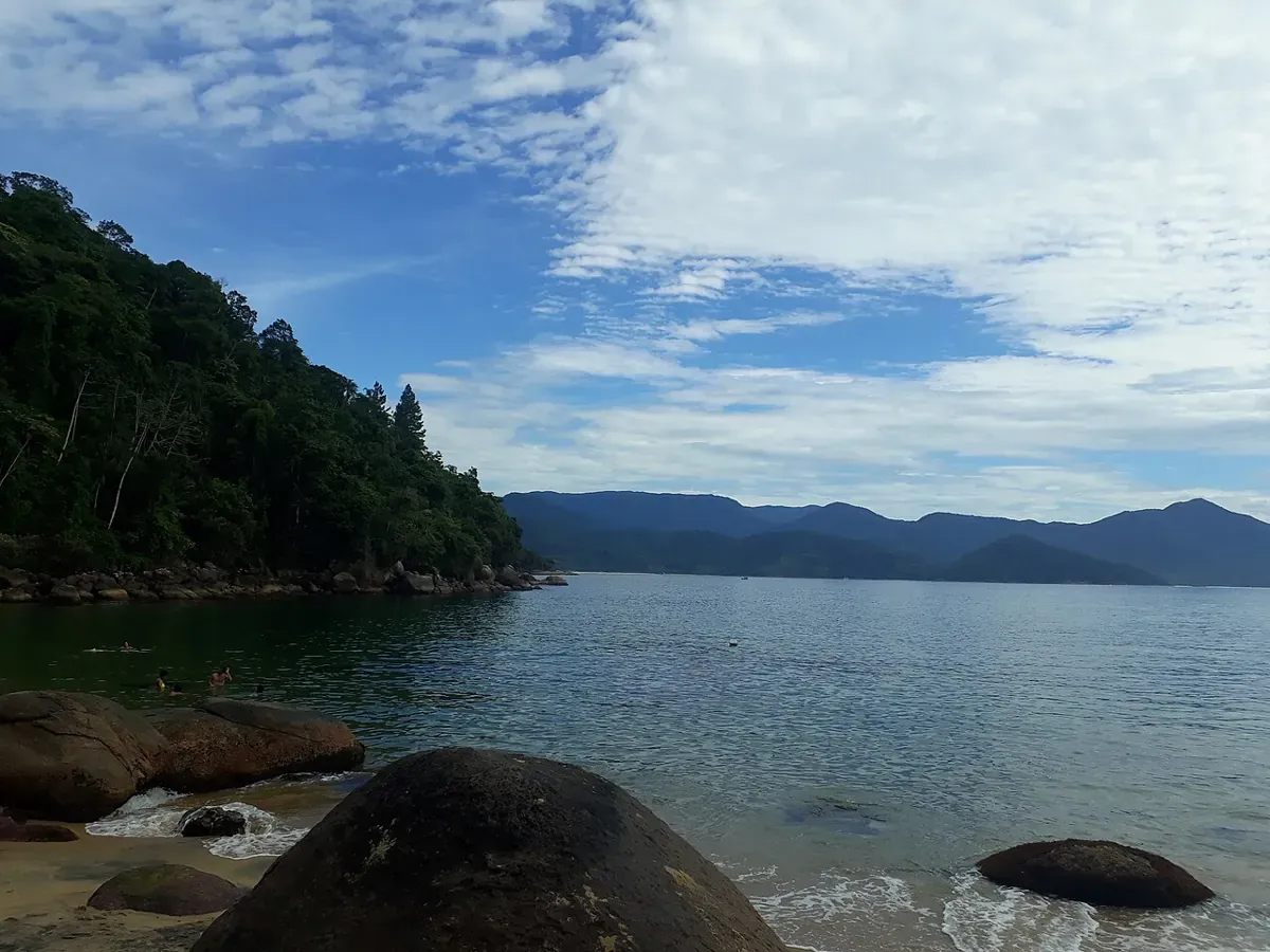Mar calmo e pedras na Praia do Cedro, com morros ao fundo